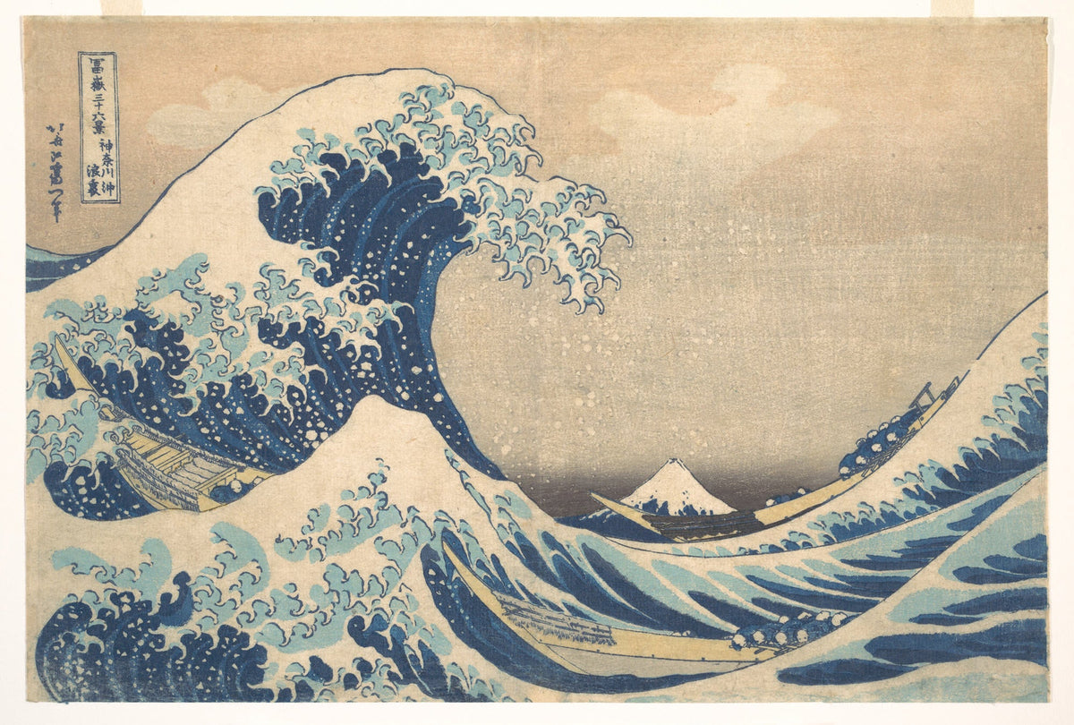 A curling blue wave arches above three narrow boats, with small Mount Fuji in the distance, in Hokusai’s woodblock print.